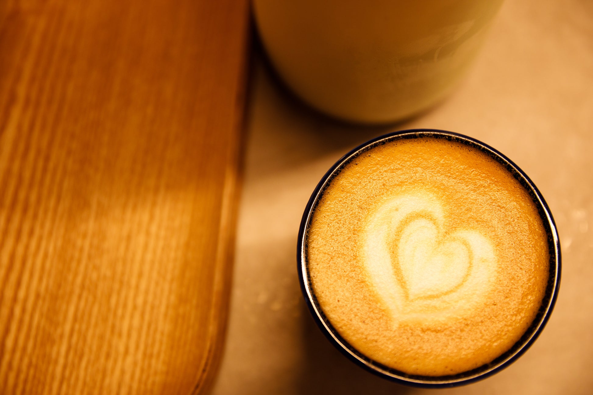 Love-shaped design on a latte, a wooden board to the left and a milk jar.