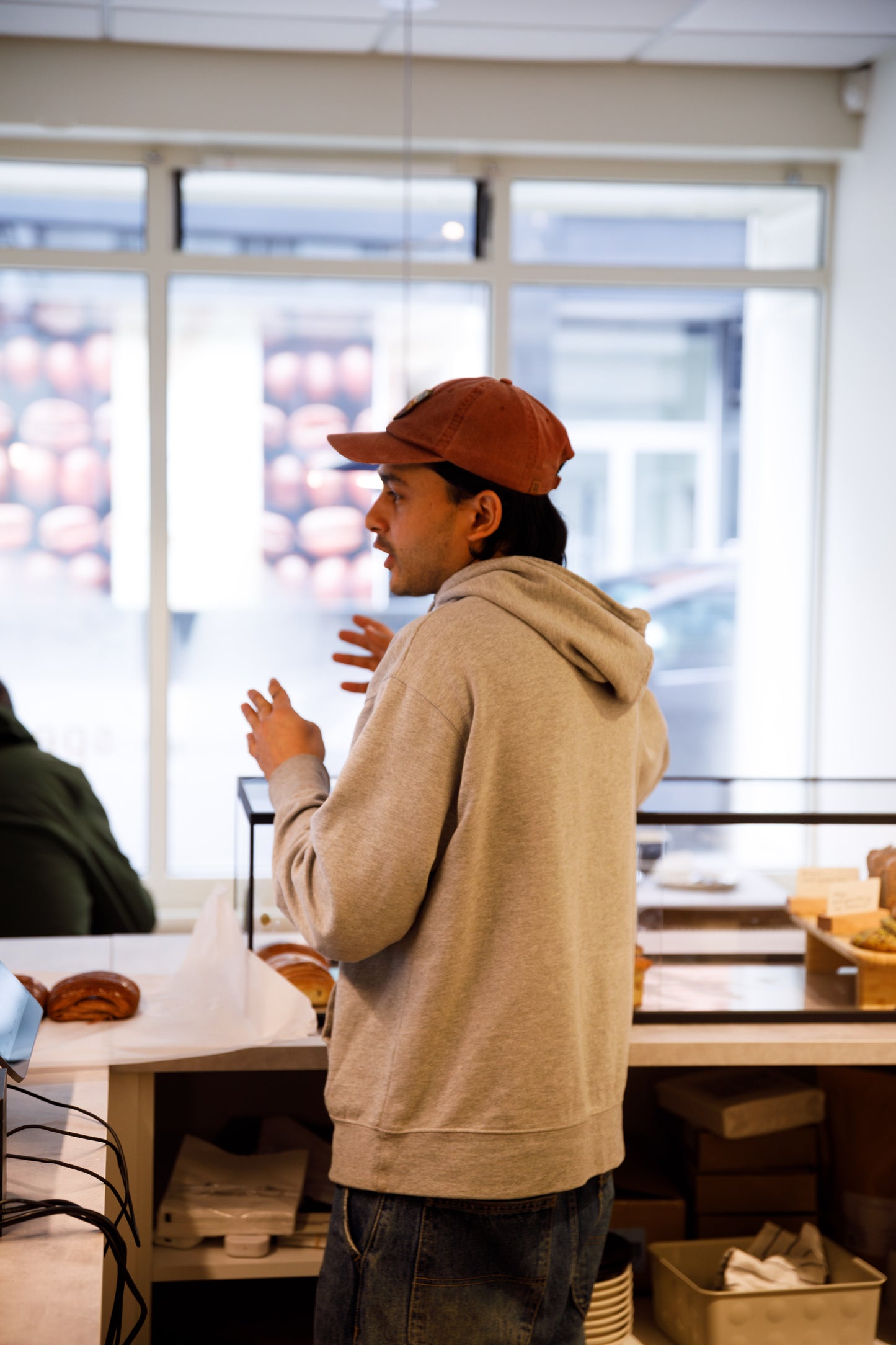 Barista mid conversation. Behind counter, fresh produce in background. Wering gry jumper, burnt orange cap.