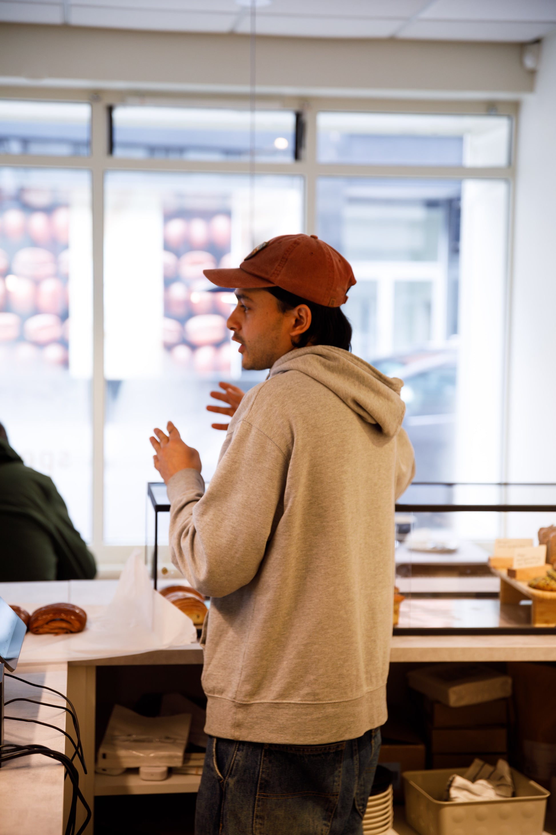 Barista mid conversation. Behind counter, fresh produce in background. Wering gry jumper, burnt orange cap.