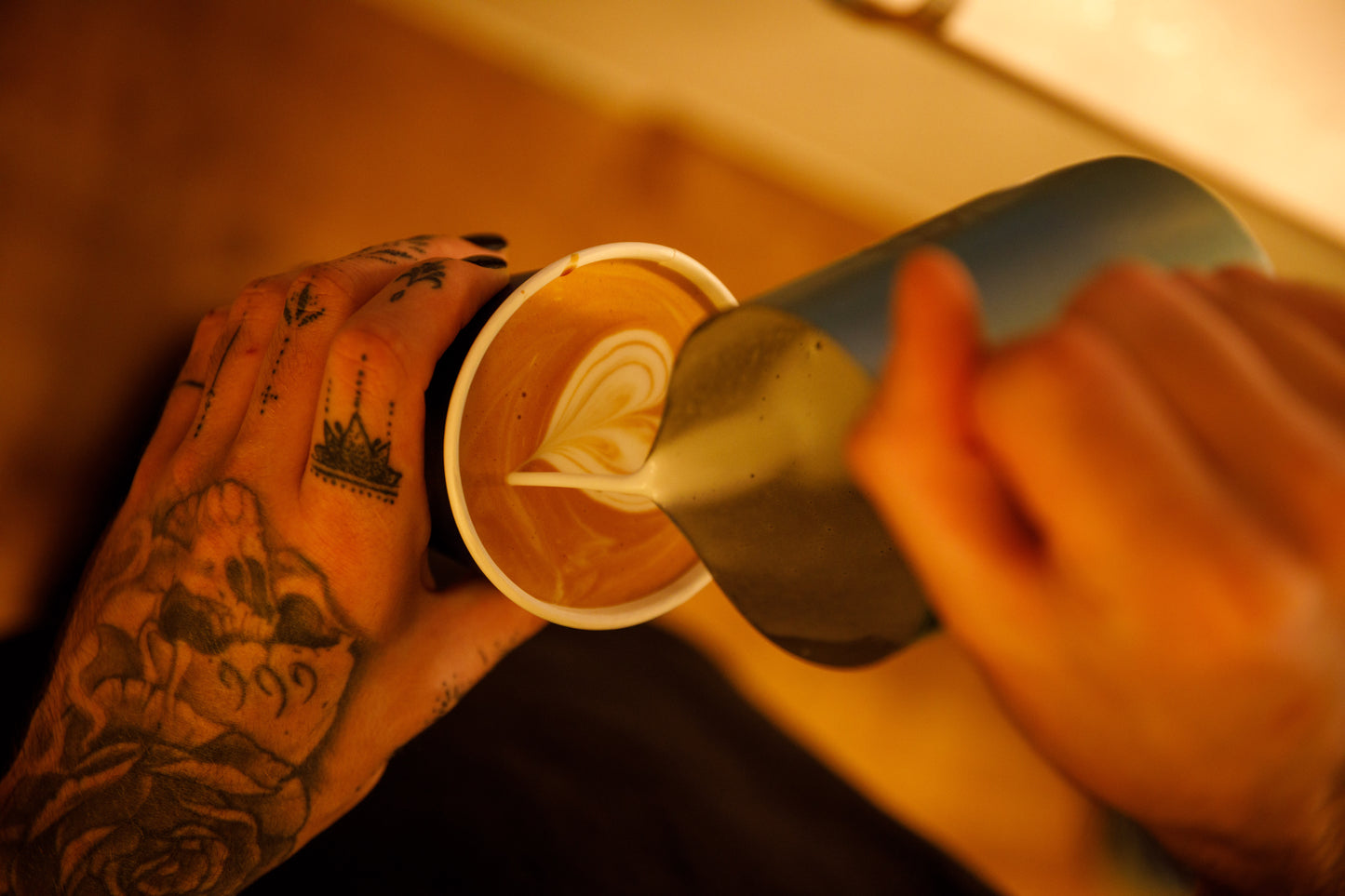 Barista pouring a milk jug with into a heart shaped design. Person has tatoos on hand.