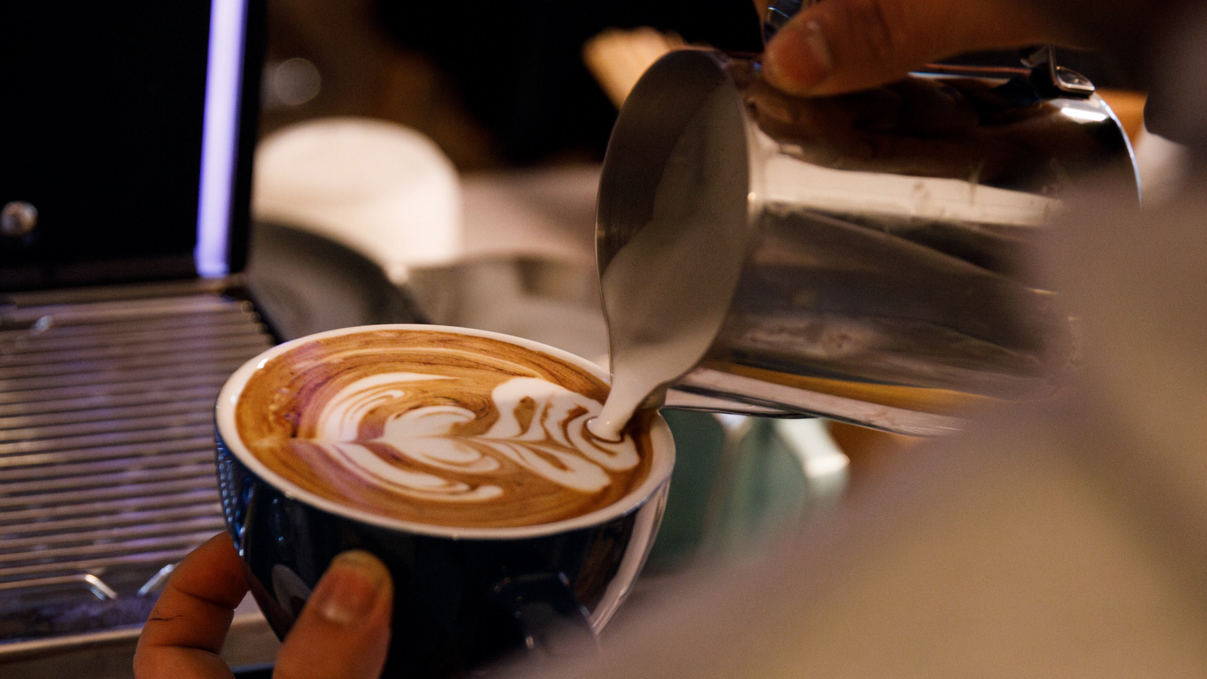 Barista designs a tulip shaped latte art in a cup of coffee. Milk jug and coffee cup in hands