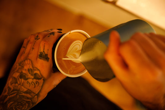Barista pouring a milk jug with into a heart shaped design. Person has tatoos on hand.
